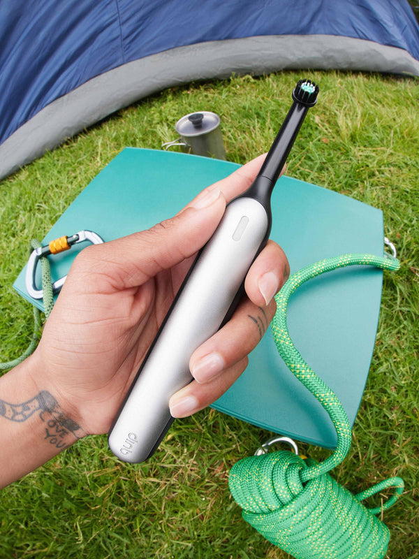 A model holds a quip 360 oscillating toothbrush in their hand, among a camping setting. 